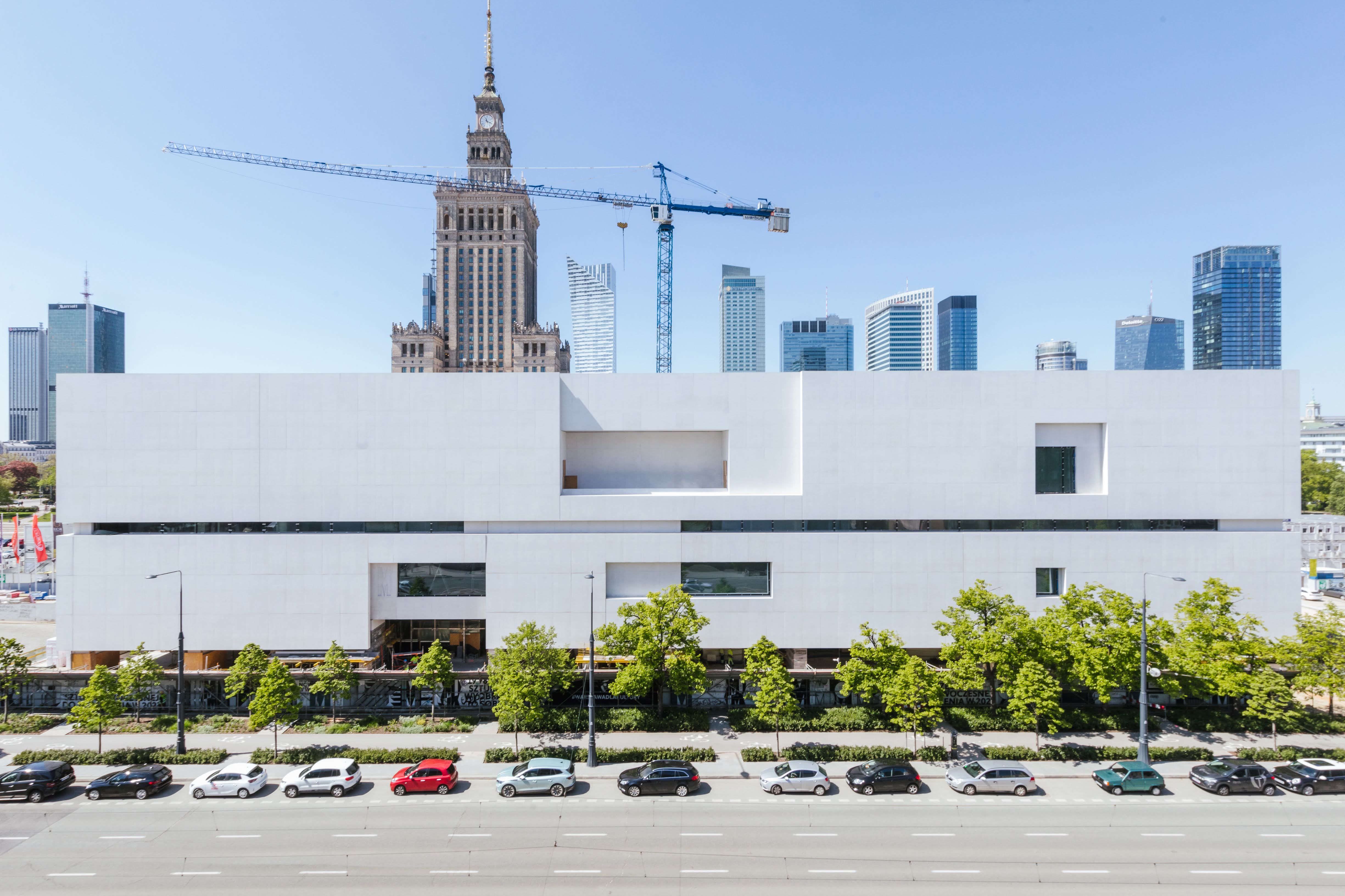 Budynek Muzeum Sztuki Nowoczesnej w Warszawie w trakcie budowy. Minimalistyczna, biała fasada z prostokątnymi otworami okiennymi. Na pierwszym planie drzewa i zaparkowane samochody wzdłuż ulicy. W tle Pałac Kultury i Nauki oraz nowoczesne wieżowce Warszawy. Widoczne dźwigi budowlane.